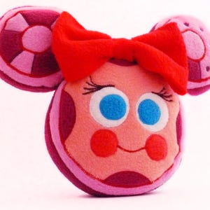 May include: A soft toy resembling a cartoon character with a round face, blue eyes, and a red bow. The toy features pink and purple details, including circular ear-like shapes. The face has a smiling expression with red cheeks.