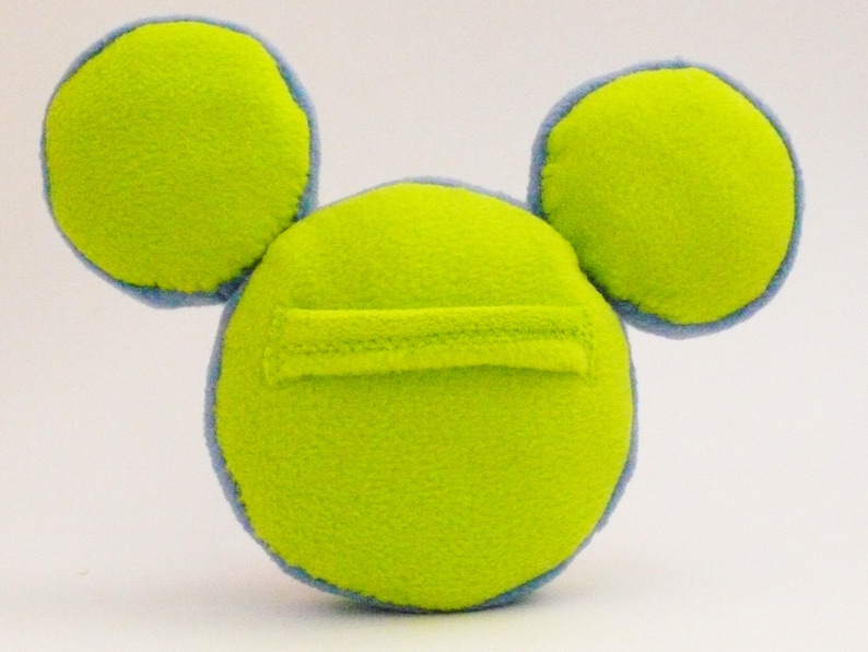 May include: A lime green plush toy with two circular ears and a circular body. The ears and body are outlined in light blue. A rectangular pocket is sewn onto the front of the body. The toy is made of soft fleece material.