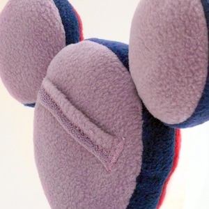 May include: A lavender-coloured ear warmer with a soft, textured surface. The ear warmer has a dark blue and red trim. The design features three rounded shapes, suggesting a playful, whimsical aesthetic. The ear warmer is designed to keep ears warm.