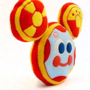 May include: A soft toy resembling a cartoon character, with a round body and large, circular ears. The toy has a light blue face with blue eyes, red cheeks, and a smiling mouth. The body and ears are red and yellow.