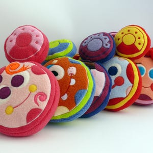 May include: A selection of brightly coloured, circular plush toys with embroidered designs. The toys display a variety of colours, such as pink, red, orange, blue, and yellow. Some have faces and decorative elements, creating a playful and whimsical appearance.