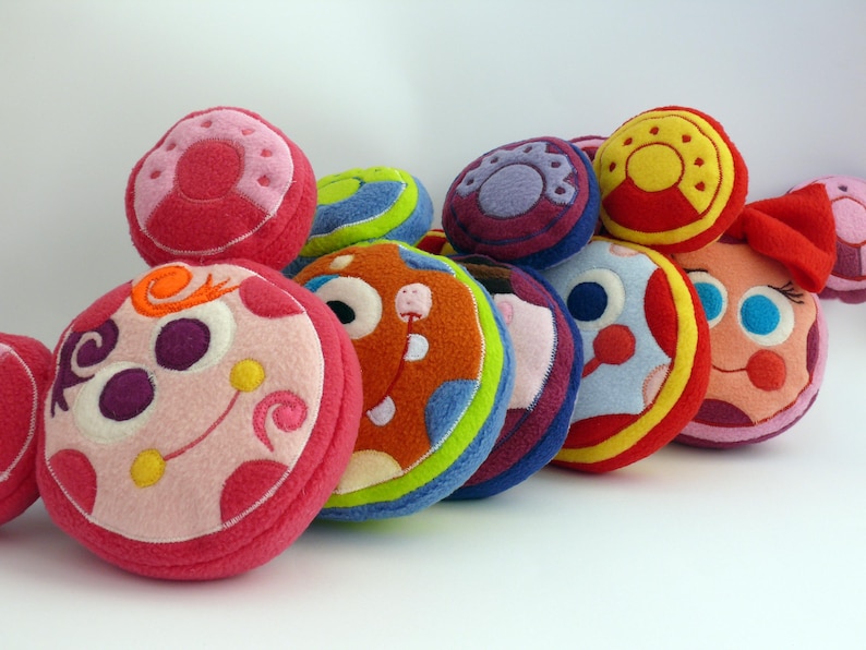 May include: A selection of brightly coloured, circular plush toys with embroidered designs. The toys display a variety of colours, such as pink, red, orange, blue, and yellow. Some have faces and decorative elements, creating a playful and whimsical appearance.