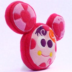 May include: A pink and white plush toy with large, round ears and a smiling face. The toy has a circular design on the ears and a whimsical expression, with purple and orange accents. The toy is designed to look like a cartoon character.