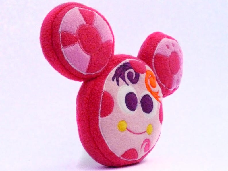 May include: A pink and white plush toy with large, round ears and a smiling face. The toy has a circular design on the ears and a whimsical expression, with purple and orange accents. The toy is designed to look like a cartoon character.
