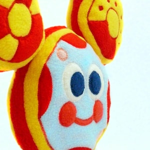 May include: A vibrant plush toy with a cartoon face and circular elements. The toy has a light blue face with large blue eyes, red cheeks, and a red smile. The body is striped with red and yellow, and the ears are yellow with red accents.