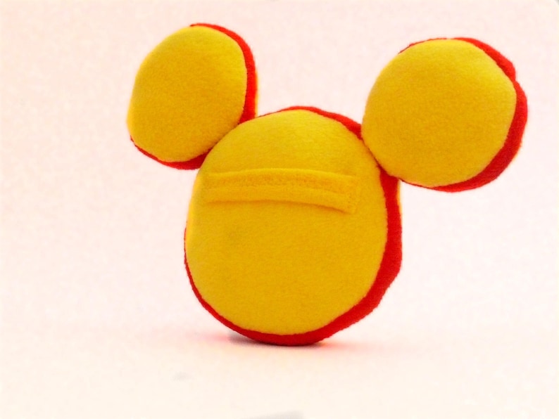 May include: A handmade plush toy in the shape of a character's head, featuring three yellow circles with red trim. The central circle has a yellow rectangular detail. The toy is made of soft fabric and is likely a children's item.
