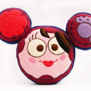 May include: A cartoon character cushion with a round face, large eyes, and a red mouth. The cushion has two circular ear-like shapes on top, one with a flower design and the other with a gear design. The color scheme includes red, pink, and blue.