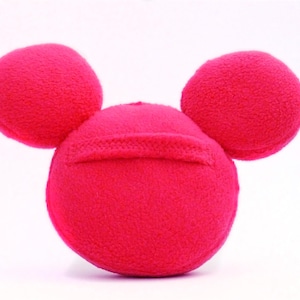 May include: A vibrant pink plush toy in the shape of a mouse's head, with a round head and two circular ears. The toy has a soft, textured surface and is set against a white background.