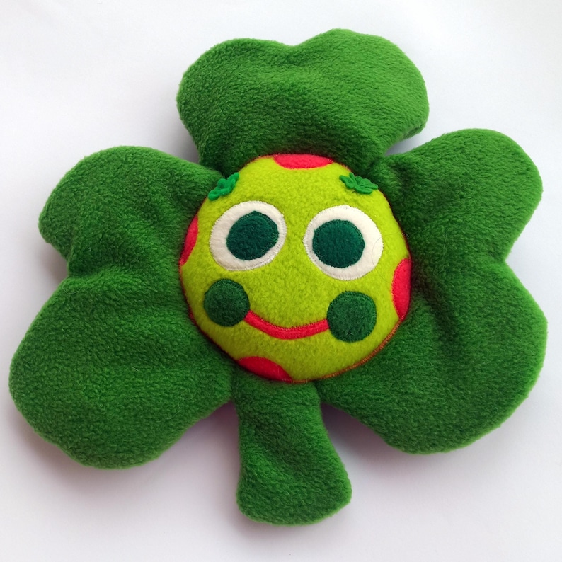 May include: A soft, green four-leaf clover toy with a smiling face. The face is lime green with red spots, large white-rimmed eyes, and a red stitched smile. The clover leaves are a darker shade of green.
