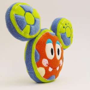 May include: A colourful plush toy shaped like a cartoon monster. The toy has an orange body with white eyes, a smiling mouth, and pink spots. The ears are blue and green. The toy is made of soft fabric.