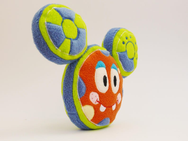 May include: A colourful plush toy shaped like a cartoon monster. The toy has an orange body with white eyes, a smiling mouth, and pink spots. The ears are blue and green. The toy is made of soft fabric.