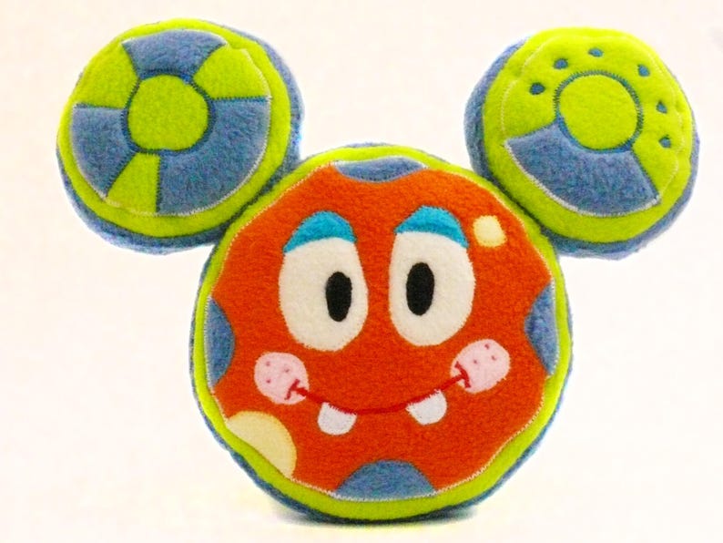 May include: A soft toy monster with a round orange body, blue and white spots, and blue eyes. It has a smiling mouth with two white teeth and two circular ears in blue and green. The toy is designed to look like a cartoon character.