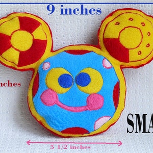 May include: A small, colorful felt character with a blue face, pink cheeks, and large yellow ears with red and yellow accents. The character is 9 inches wide, 7 inches tall, and 5.5 inches across. The word "SMALL" is printed in black.
