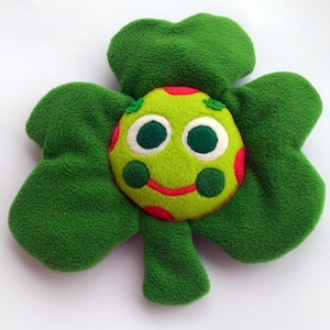 May include: A green, flower-shaped soft toy with a smiling face at the centre. The face is lime green with red spots, large black eyes, and a red smile. The flower has four green petals.
