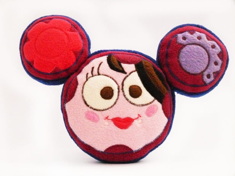 May include: A plush, cartoon-style character pillow with a round face, large eyes, and a red smiling mouth. The pillow has two circular ear-like shapes on top, one with a flower design and the other with a gear design. The color scheme includes red, pink, and blue.