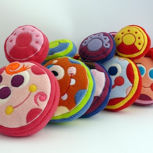 May include: A selection of brightly coloured, round, plush toys with quirky designs. The toys display a variety of colours, such as pink, red, orange, blue, and yellow, with embroidered details and playful faces. These soft toys are likely designed for children.
