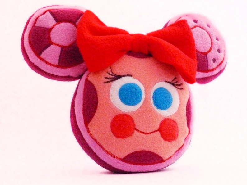 May include: A soft toy resembling a cartoon character with a round face, blue eyes, and a red bow. The toy features pink and purple details, including circular ear-like shapes. The face has a smiling expression with red cheeks.