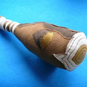May include: A handcrafted felt club toy in shades of brown, tan, and cream, with a rounded head and a wrapped handle. The head features a circular design and stitched details, ideal for imaginative play.