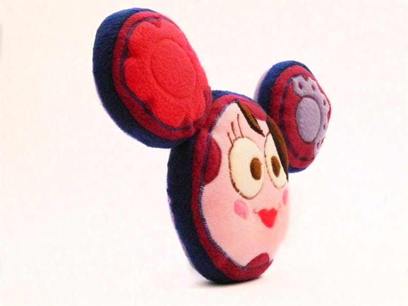 May include: A plush doll with a cartoon face, featuring large, round eyes, a red heart-shaped mouth, and rosy cheeks. The doll has large, rounded ears in red and blue, and a small, purple paw print on one ear. The doll is made of soft fabric.