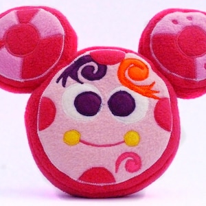May include: A soft toy resembling a cartoon character's face, with large purple eyes, a smiling mouth, and two circular ears. The toy is mainly pink with lighter pink and orange details.