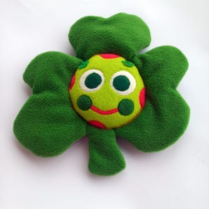 May include: A plush green shamrock-shaped toy with a smiling face. The face is light green with red polka dots, large white-rimmed eyes, and a red smiling mouth. The shamrock has four large, soft, green leaves.