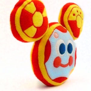 May include: A soft toy resembling a cartoon character, with a round body and large, circular ears. The toy has a light blue face with blue eyes, red cheeks, and a smiling mouth. The body and ears are red and yellow.