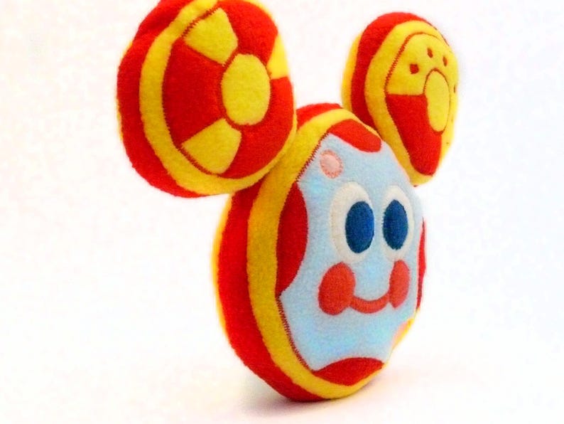 May include: A soft toy resembling a cartoon character, with a round body and large, circular ears. The toy has a light blue face with blue eyes, red cheeks, and a smiling mouth. The body and ears are red and yellow.