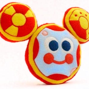 May include: A soft toy shaped like a cartoon character's head, with a light blue face, large blue eyes, red cheeks, and a smiling mouth. It has two circular ears in red and yellow. The toy is made from soft fabric.