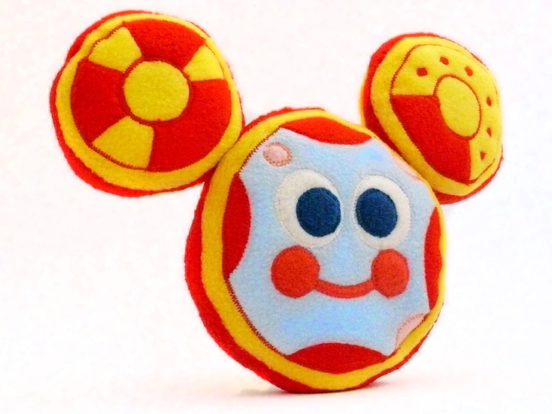 May include: A soft toy shaped like a cartoon character's head, with a light blue face, large blue eyes, red cheeks, and a smiling mouth. It has two circular ears in red and yellow. The toy is made from soft fabric.