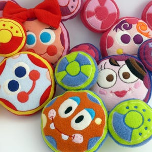 May include: A selection of colourful, plush, round toys with embroidered faces and designs. The toys feature various colours, including red, yellow, blue, green, and orange. Some have bow accents, while others have circular patterns and facial features.