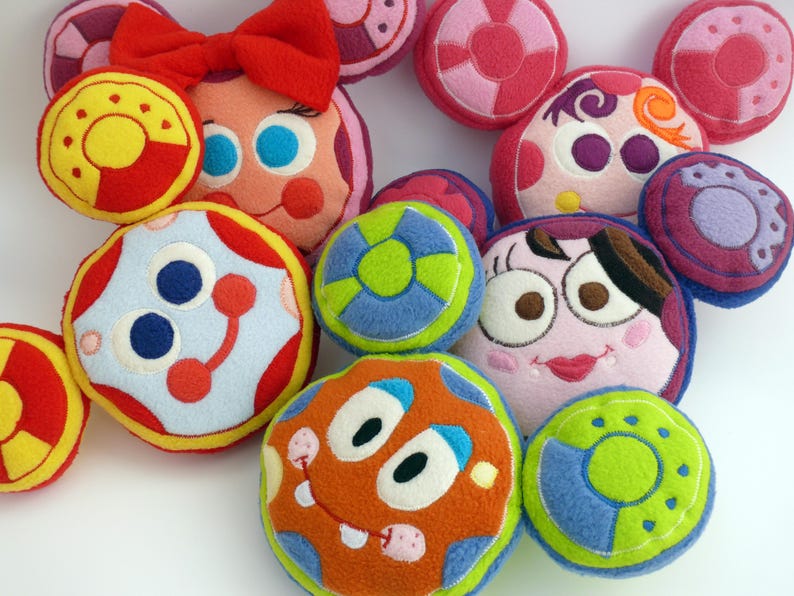 May include: A selection of colourful, plush, round toys with embroidered faces and designs. The toys feature various colours, including red, yellow, blue, green, and orange. Some have bow accents, while others have circular patterns and facial features.