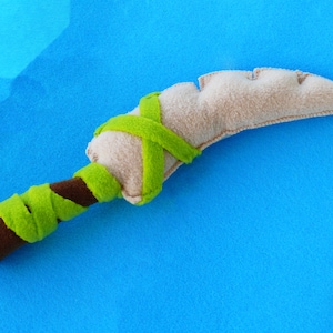 Peut inclure: Un poignard en peluche avec une lame beige et un manche marron enveloppé de tissu vert. La lame a un bord dentelé et le manche est cylindrique. Le jouet est posé sur un fond bleu vif.