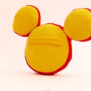 May include: A handmade yellow plush toy with red trim, featuring a circular body and two circular ears. The toy has a small yellow rectangular detail on the body. The toy is made of soft fabric.