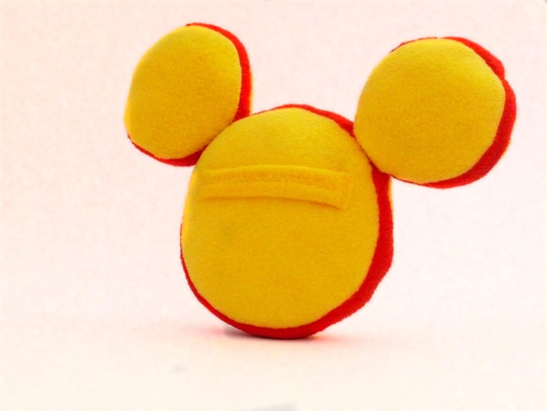 May include: A handmade yellow plush toy with red trim, featuring a circular body and two circular ears. The toy has a small yellow rectangular detail on the body. The toy is made of soft fabric.