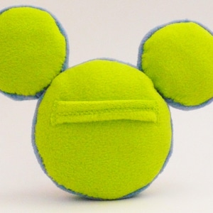 May include: A lime green plush toy with two circular ears and a circular body. The ears and body are outlined in light blue. A rectangular pocket is sewn onto the front of the body. The toy is made of a soft, fuzzy material.