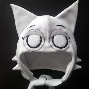 Op de afbeelding: Een witte kattenorenhoed met grote, gesloten ogen en een gestikte zwarte omtrek. De hoed heeft puntige oren en strikken onder de kin. De ogen zijn groot en rond, met zwarte gestikte contouren en witte centra.