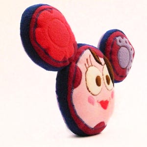 May include: A plush doll with a cartoon face, featuring large, round eyes, a red heart-shaped mouth, and rosy cheeks. The doll has large, rounded ears in red and blue, and a small, purple paw print on one ear.