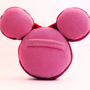 May include: A pink plush toy with a circular body and two rounded ear-like shapes on top. The toy has a stitched rectangular detail on the front and a dark red trim.