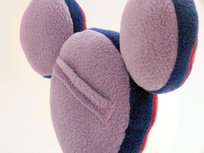 May include: A lavender-coloured ear warmer with a soft, textured surface. The ear warmer has a dark blue and red trim. The design features three rounded shapes, suggesting a playful, whimsical aesthetic. The ear warmer is designed to keep ears warm.