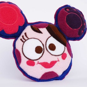 May include: A plush toy in the shape of a cartoon character's face. The toy features large, expressive eyes, a smiling mouth, and large ears. The color palette includes pink, red, purple, brown, and blue, with contrasting stitching.
