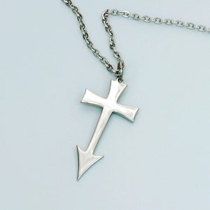 May include: A silver-colored necklace with a cross pendant. The cross has a pointed bottom and is suspended from a chain. The jewelry is displayed against a light blue background.