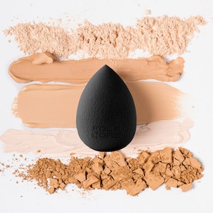 May include: A black, teardrop-shaped makeup sponge is positioned on a white background, with swatches of foundation and loose powder in beige and brown shades. The sponge has a textured surface, designed for makeup application.