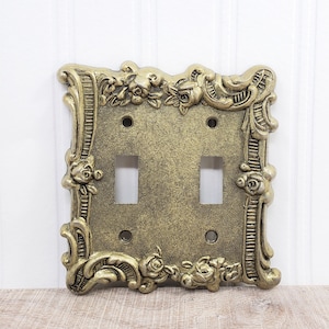 Könnte beinhalten: Verzierte, antike Doppel-Lichtschalterabdeckung in Goldoptik. Die Abdeckung hat ein dekoratives Blumen- und Scroll-Design an den Rändern, mit zwei Schalteröffnungen und Schraubenlöchern.