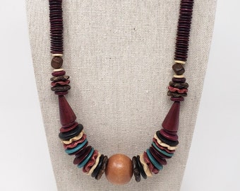 May include: A wooden necklace with a variety of shapes and colors. The necklace features a central large wooden bead, flanked by sections of red, black, blue, and cream-colored wooden discs. The necklace is displayed on a neutral-toned mannequin.