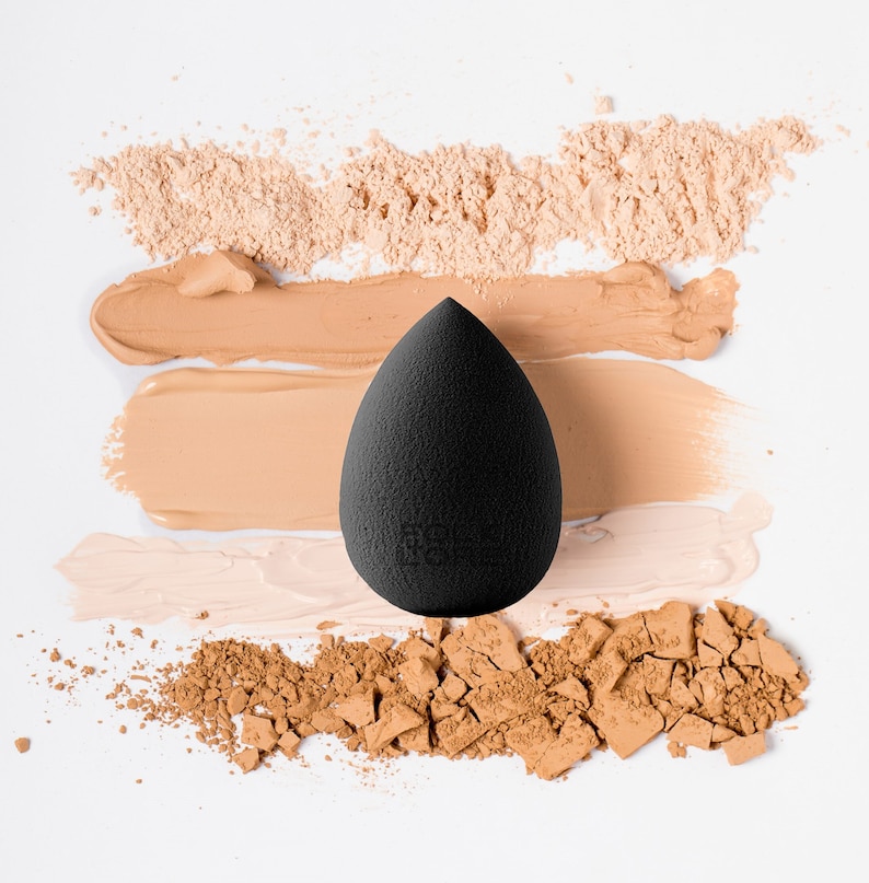 May include: A black, teardrop-shaped makeup sponge is positioned on a white background, with swatches of foundation and loose powder in beige and brown shades. The sponge has a textured surface, designed for makeup application.