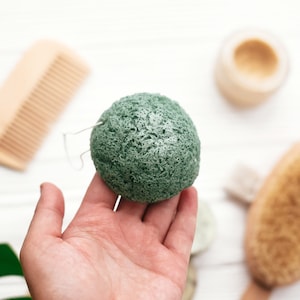 Puede incluir: Una esponja verde y redonda con un lazo de alambre, sostenida en una mano. Otros productos de baño incluyen un peine de madera, un tarro de crema y un cepillo corporal, todo sobre una superficie de madera blanca. Los artículos sugieren un enfoque en el cuidado natural de la piel.