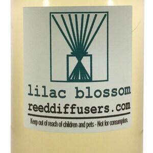 May include: A clear glass bottle with a white cap filled with a light yellow liquid. The bottle has a label with a green logo of a reed diffuser and the text "lilac blossom reeddiffusers.com". The label also includes the text "Keep out of reach of children and pets - Not for consumption."