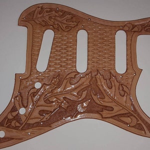 Puede incluir: Una placa de protecci&oacute;n de guitarra de cuero marr&oacute;n con un patr&oacute;n tejido y un intrincado dise&ntilde;o de hojas.