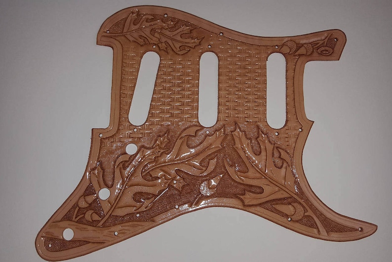 Puede incluir: Una placa de protecci&oacute;n de guitarra de cuero marr&oacute;n con un patr&oacute;n tejido y un intrincado dise&ntilde;o de hojas.