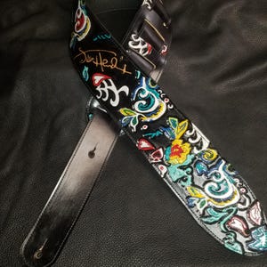 May include: A black leather guitar strap with a colorful paisley design and the signature "Jim Heath".